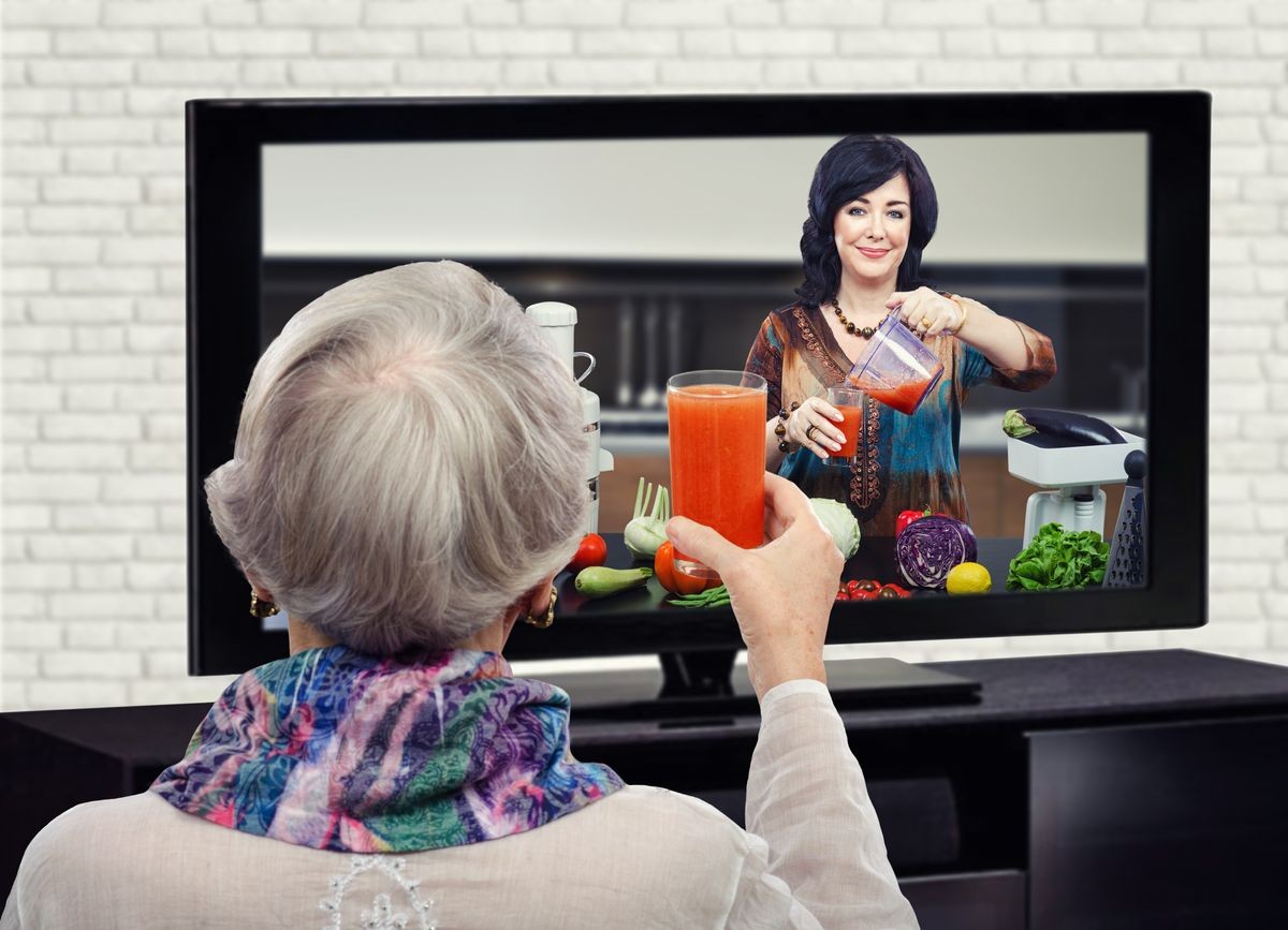 Grey haired woman raising glass of detox drink in front of TV screen. In the screen, nutrition consultant shows how to make healthy vegetables juice at home